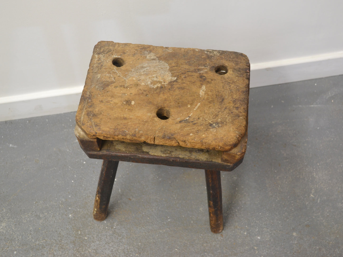 Old wooden stool with three holes on a gray floor against a white wall.